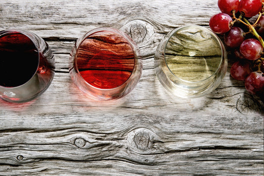 Tres copas de vino tinto, rosado y blanco se ven desde arriba sobre una mesa de madera desgastada; a un lado hay un racimo de uvas rojas.