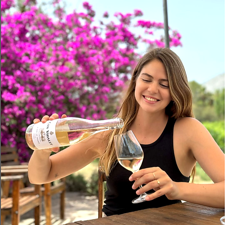 Mujer joven sonriendo mientras sirve vino blanco Can Vidalet en una copa al aire libre, rodeada de buganvillas en flor y el paisaje soleado de Mallorca. La imagen transmite la alegría, frescura y autenticidad del enoturismo mallorquín y del vino artesanal de la bodega Can Vidalet.