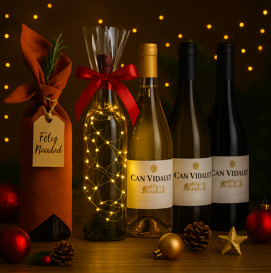 Cinco botellas de vino Can Vidalet en una mesa decorada para Navidad; una botella envuelta en tela estilo furoshiki con una etiqueta “Feliz Navidad”, otra decorada con luces LED, y tres botellas de vino. El fondo presenta luces cálidas y adornos festivos.