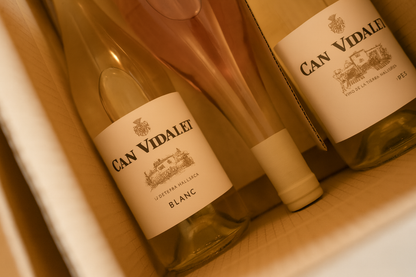 Tres botellas de vino de Bodegas Can Vidalet dentro de una caja de cartón color kraft. A la izquierda y derecha se aprecian vinos blancos, y en el centro una botella de vino rosado. Las etiquetas blancas muestran el logotipo y la ilustración de la finca Can Vidalet, destacando la producción artesanal de vino mallorquín. La imagen transmite elegancia, autenticidad y el cuidado en la presentación de los vinos seleccionados.