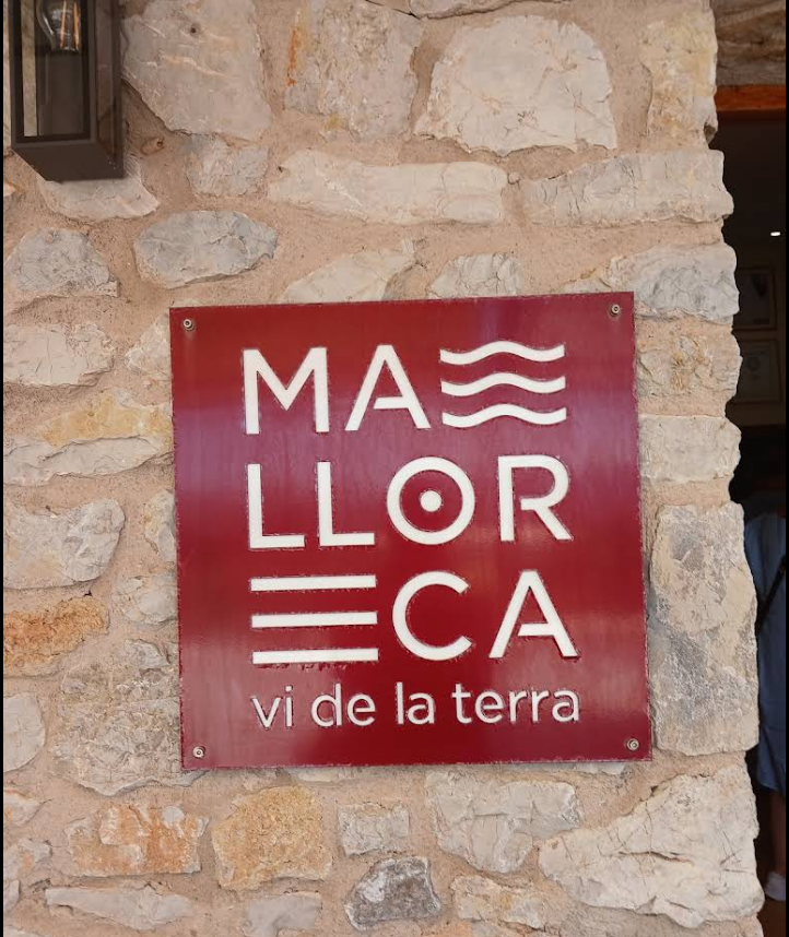 Cartel rojo con letras blancas que dice “Mallorca – Vi de la Terra”, fijado sobre una pared de piedra típica mallorquina. Este distintivo representa la indicación geográfica protegida de los vinos elaborados en la isla de Mallorca, símbolo de calidad, origen local y tradición vitivinícola balear.