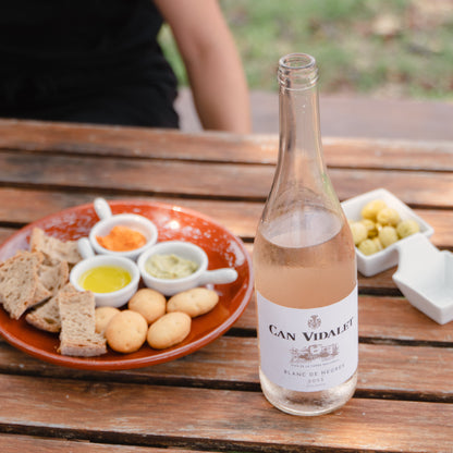 Mesa de cata al aire libre en Bodegas Can Vidalet, Mallorca. En la mesa de madera se muestra una botella de vino rosado Can Vidalet Blanc de Negres 2023 junto a un plato con tapas veganas: pan artesanal, aceitunas verdes, panecillos, aceite de oliva virgen extra y salsas mediterráneas de pimientos y hierbas. Una persona disfruta del ambiente relajado de la bodega entre naturaleza y vino mallorquín artesanal.