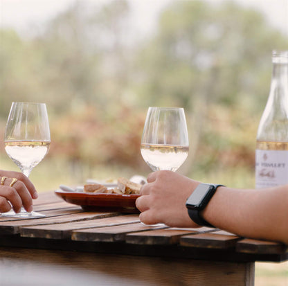 Dos personas disfrutan de copas de vino blanco en una mesa de madera al aire libre, con una botella de Can Vidalet y un paisaje natural desenfocado al fondo.